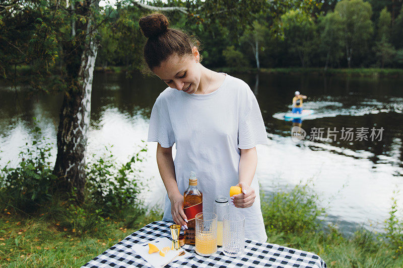 Happy female making cocktails by the lake during summer picnic